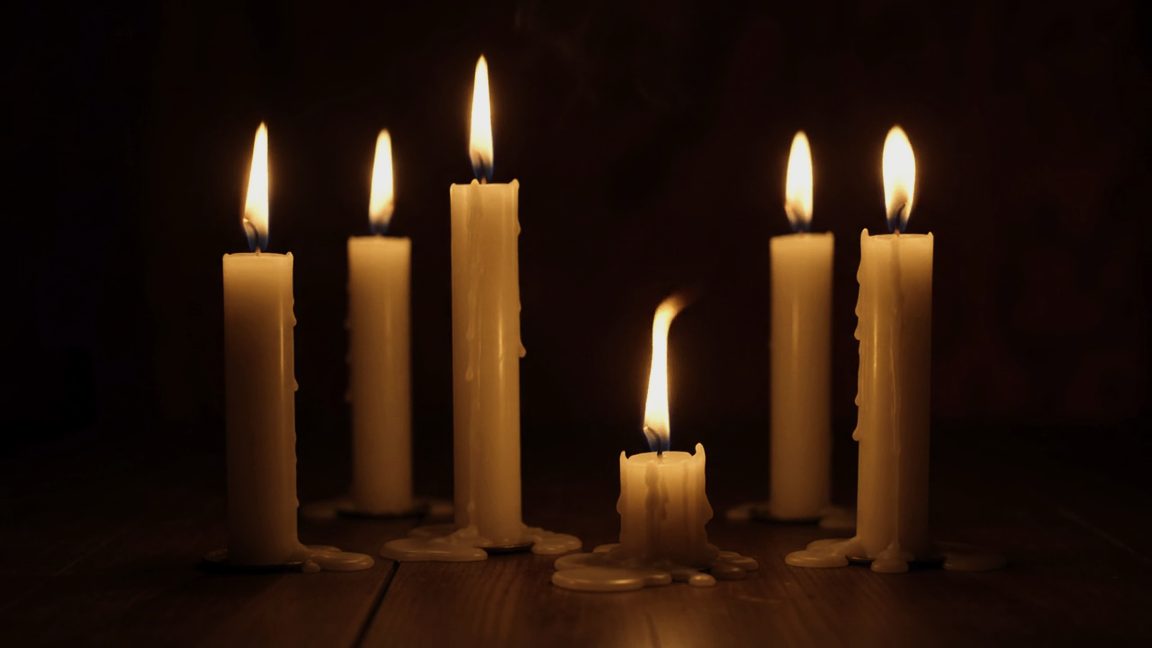 Six candles burning on a dark table, five tall and one burned almost to the wood, its flame guttering.